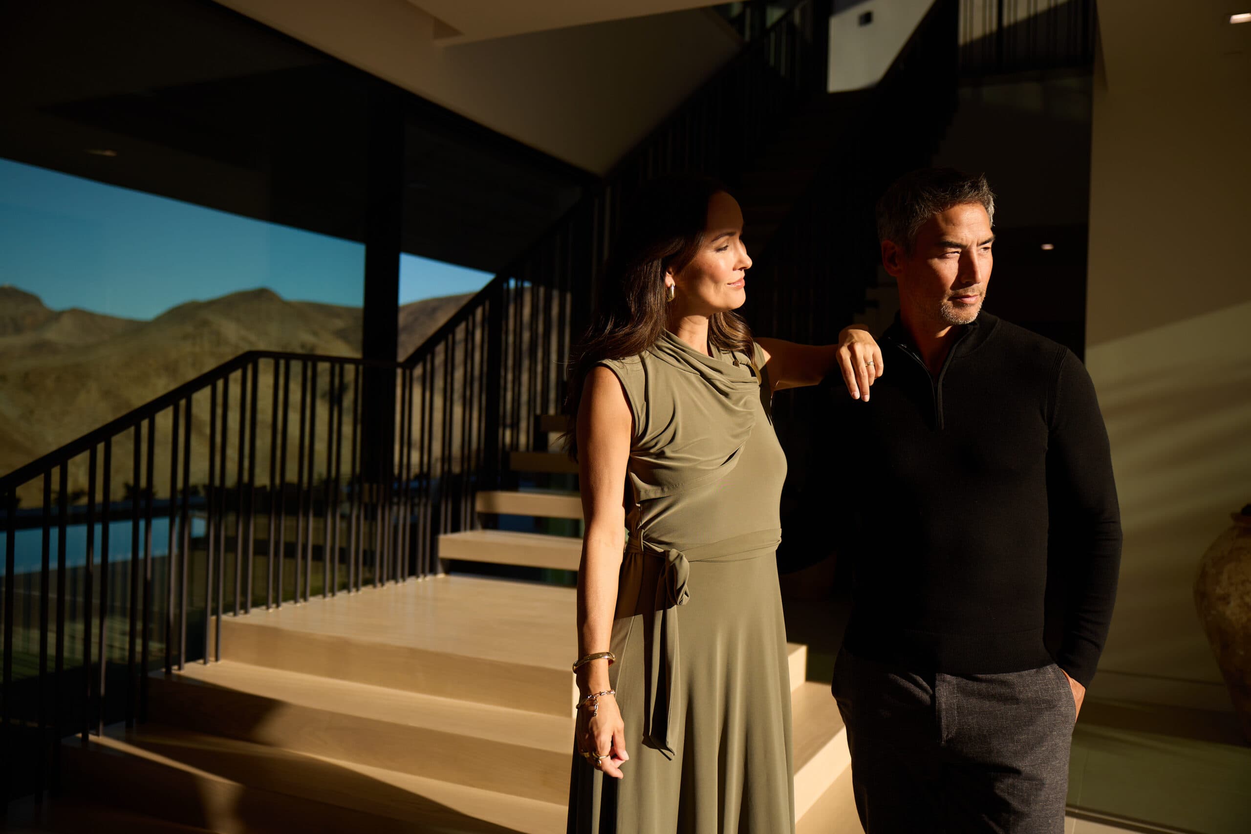 Couple standing in a modern luxury home interior with floor-to-ceiling windows, warm sunlight, and mountain views, showcasing upscale contemporary living.