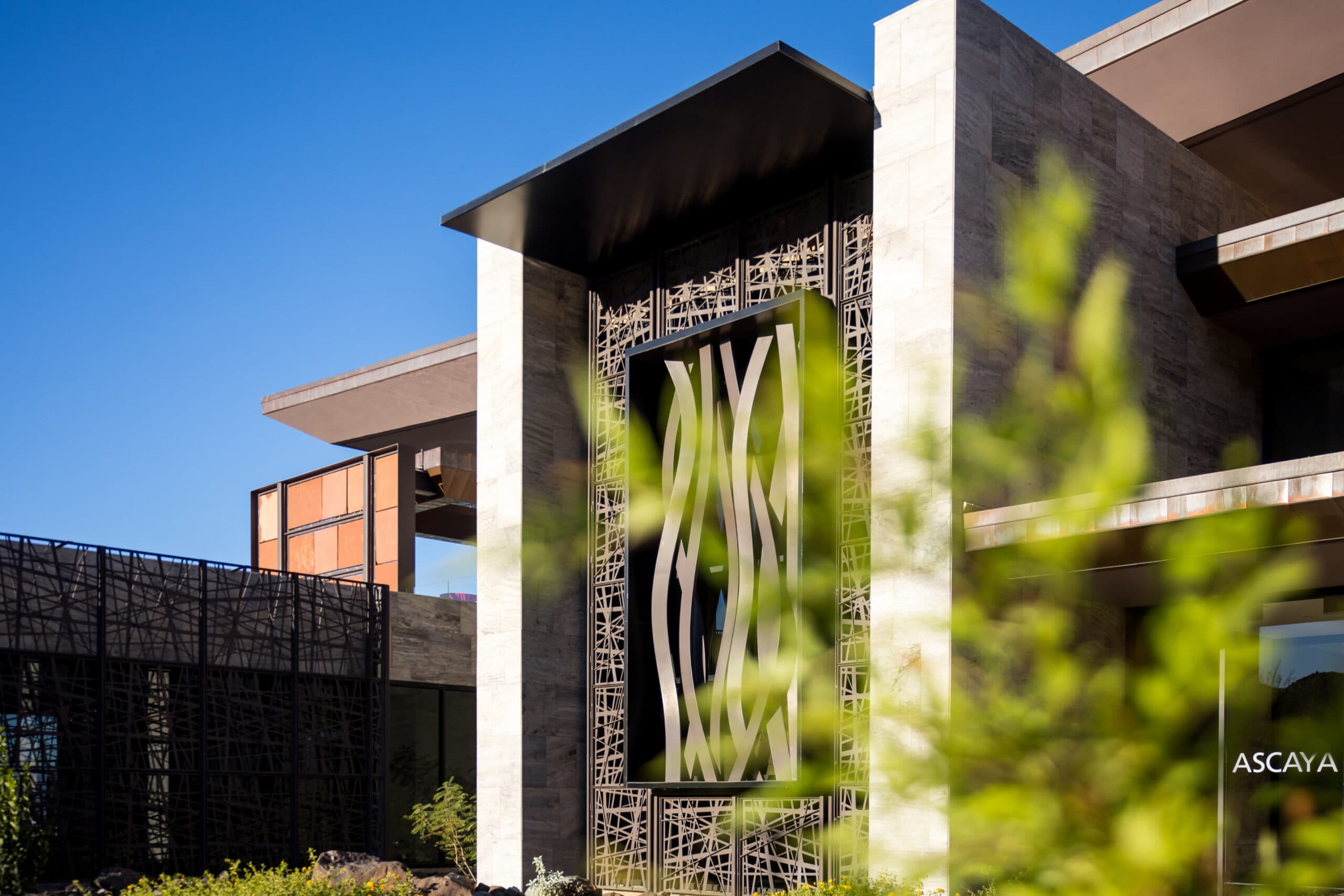 Modern architectural entrance at Ascaya featuring stone walls, geometric metal art installation, and landscaped greenery under a clear blue sky.