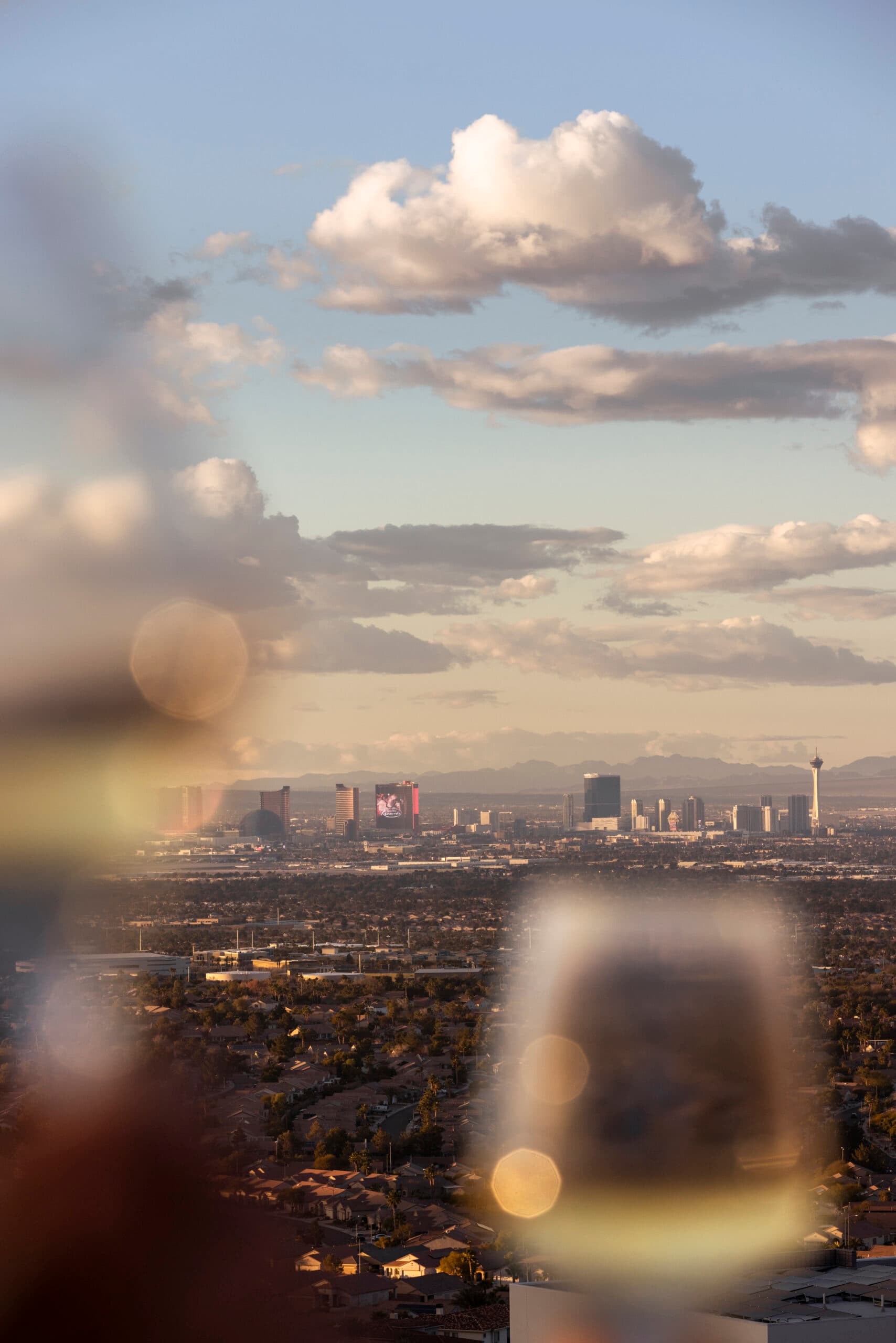 Distant view of the Las Vegas skyline at sunset with soft clouds overhead, seen from a residential area with warm light and blurred foreground elements.