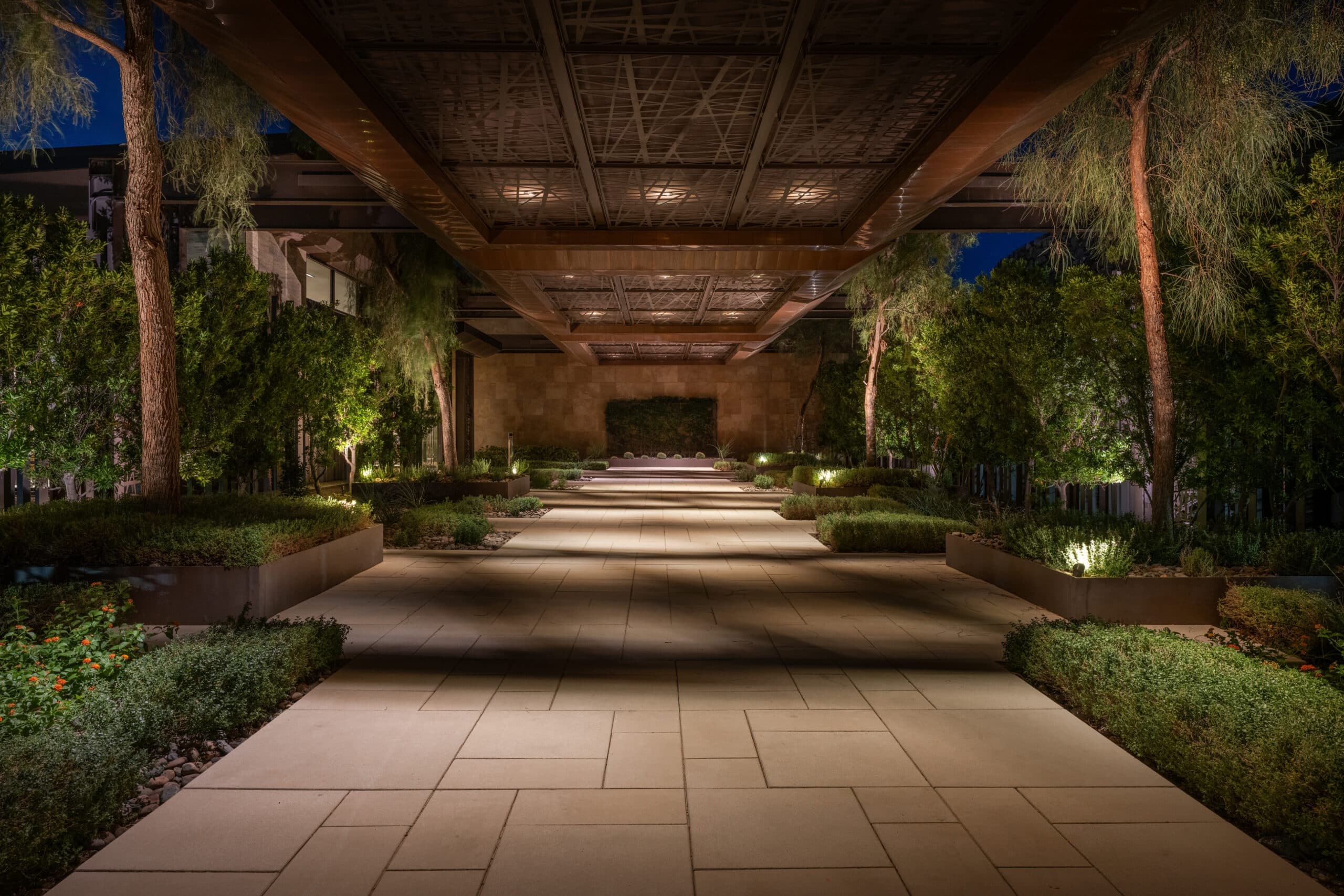 A symmetrical, modern outdoor walkway at night with warm ambient lighting, framed by trees and landscaped greenery, beneath a large architectural canopy with geometric detailing.